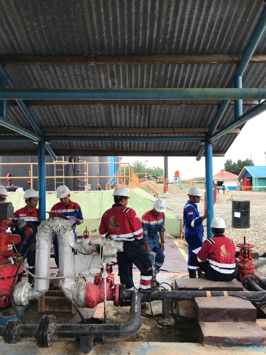Tim CIK CORP mengerjakan manifold sumur di bawah kanopi, dengan tangki dan fasilitas produksi di latar belakang